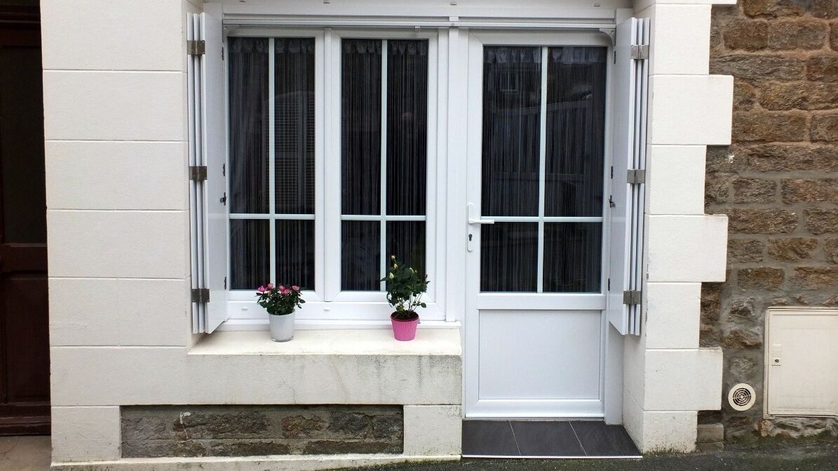 A building with a white facade, a stone wall on the right, and a window with a white frame and a door.