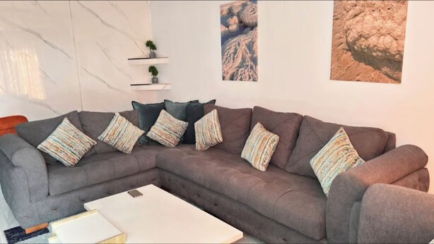 Een moderne woonkamer met een grijze hoekbank, een witte salontafel en decoratieve kussens. Er zijn wandplanken met planten en een groot kunstwerk in een lijst.