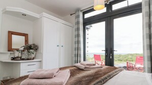 Een slaapkamer met een groot bed, een zacht tapijt, een nachtkastje met een vaas en glazen, en uitzicht op het groen door een groot raam.