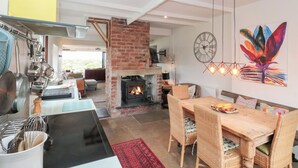 Een moderne keuken met een bakstenen open haard, een eethoek met rieten stoelen, en een kleurrijk kunstwerk aan de muur.