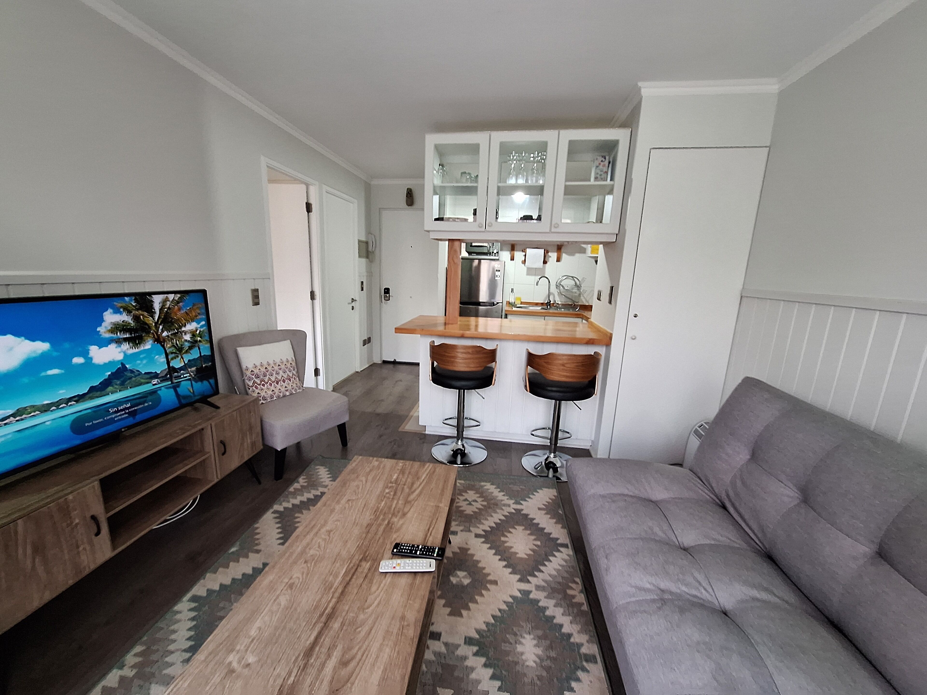 Un espacio habitable compacto con televisor, sofá, mesa de centro y una zona de cocina con taburetes de bar.