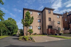 A multi-story building with a brick facade, a central entrance, and a small parking area with trees. - The Well - 3 bedroom apartment (Motherwell)