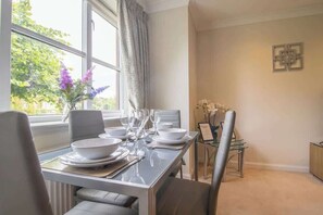 A dining area with a table set for four, chairs, a vase with flowers, and a window with curtains. - The Well - 3 bedroom apartment (Motherwell)