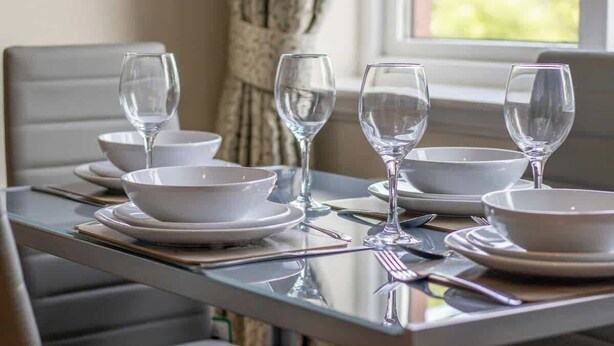 A dining table set with white plates, bowls, and wine glasses, with a window in the background. - The Well - 3 bedroom apartment (Motherwell)