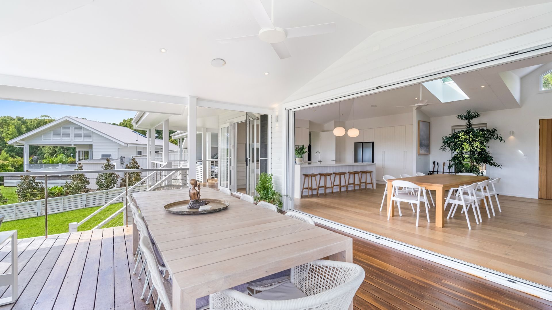 A spacious dining area with a wooden table and white chairs, a kitchen with white cabinets, and a view of a house and greenery outside.