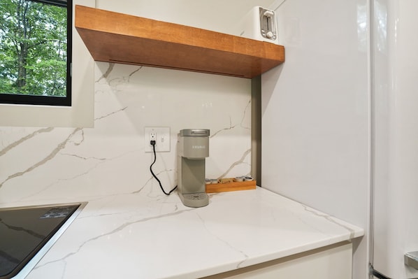 A kitchen with a marble countertop, a Keurig coffee maker, a wooden shelf, and a window with a view of trees.