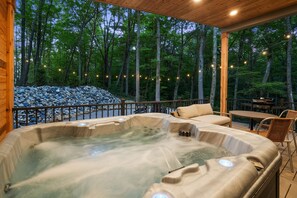A hot tub area with a wooden deck, surrounded by a stone wall and trees, and string lights.