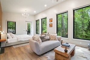 A modern bedroom with a large bed, a grey sofa, and a wooden coffee table. There are large windows with a forest view.