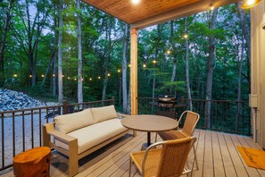 A wooden deck with a sofa and chairs, a grill, and a view of a forested area.