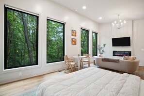A modern bedroom with large windows, a flat-screen TV, a fireplace, and a cozy seating area.