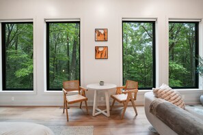 A modern living room with large windows, a round table, two wooden chairs, and a sofa.