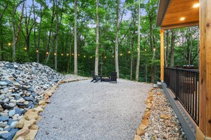 A covered outdoor seating area with a fire pit, surrounded by a gravel path and a rock border, set in a forested area with string lights. - Cliffside View | Modern Couples Retreat, Hot Tub (Rockbridge)