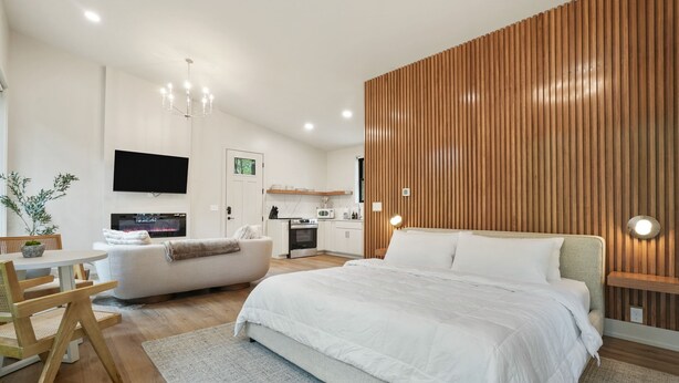 Un dormitorio moderno con una cama grande, un televisor montado en la pared, un revestimiento de pared de madera y una pequeña kitchenette al fondo.