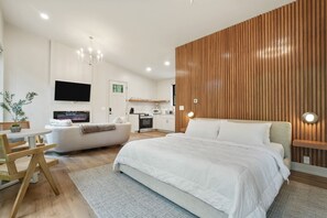 A modern bedroom with a large bed, a TV mounted on the wall, a wooden accent wall, and a small kitchenette in the background. - Cliffside View | Modern Couples Retreat, Hot Tub (Rockbridge)