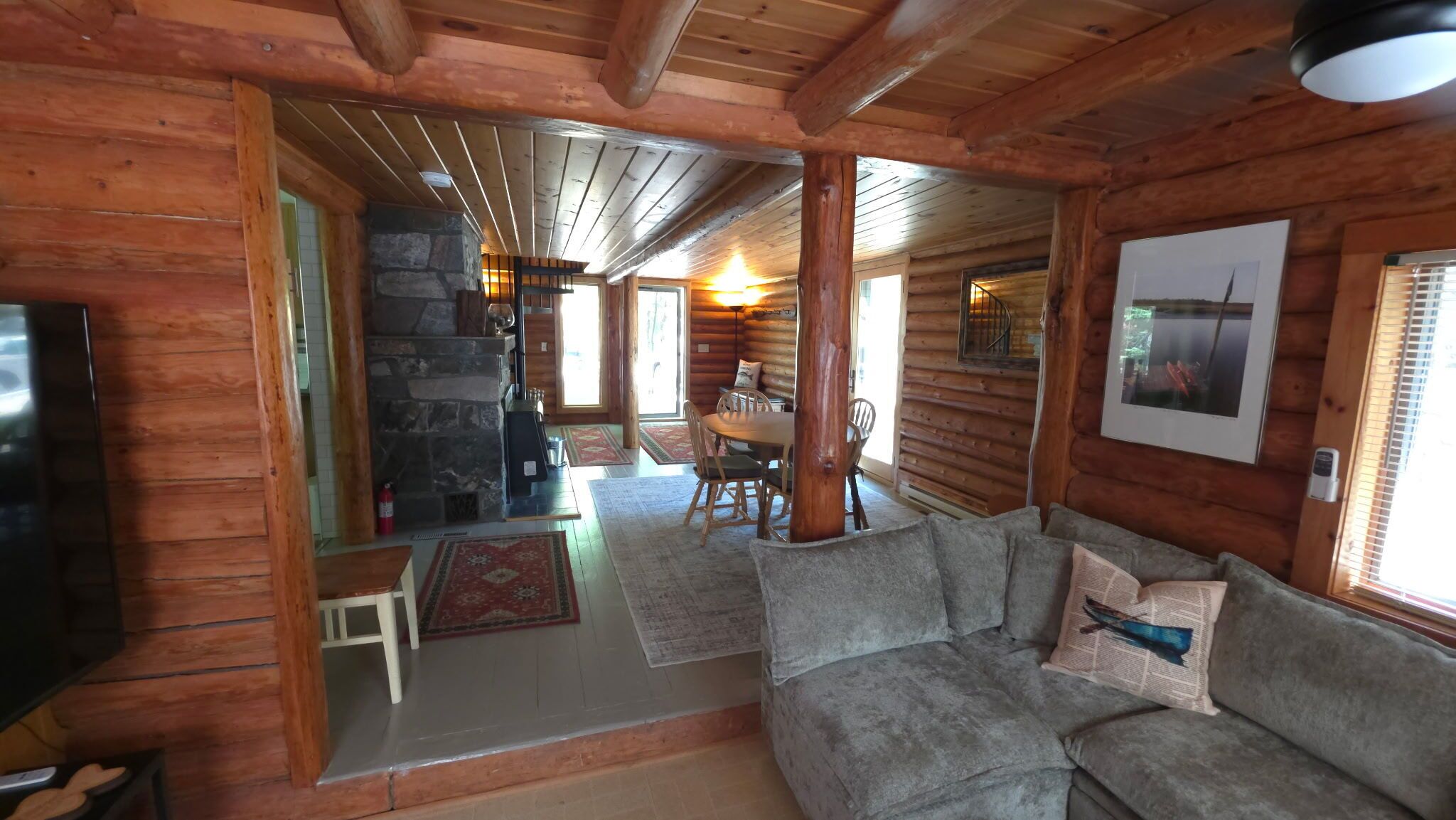 A log cabin interior with a stone fireplace, wooden dining table, and a gray sofa.