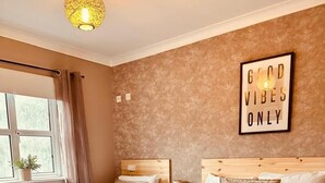 Habitación de hotel con dos camas, una ventana con cortinas, un cartel en la pared que dice "GOOD VIBES ONLY" y una lámpara colgante.