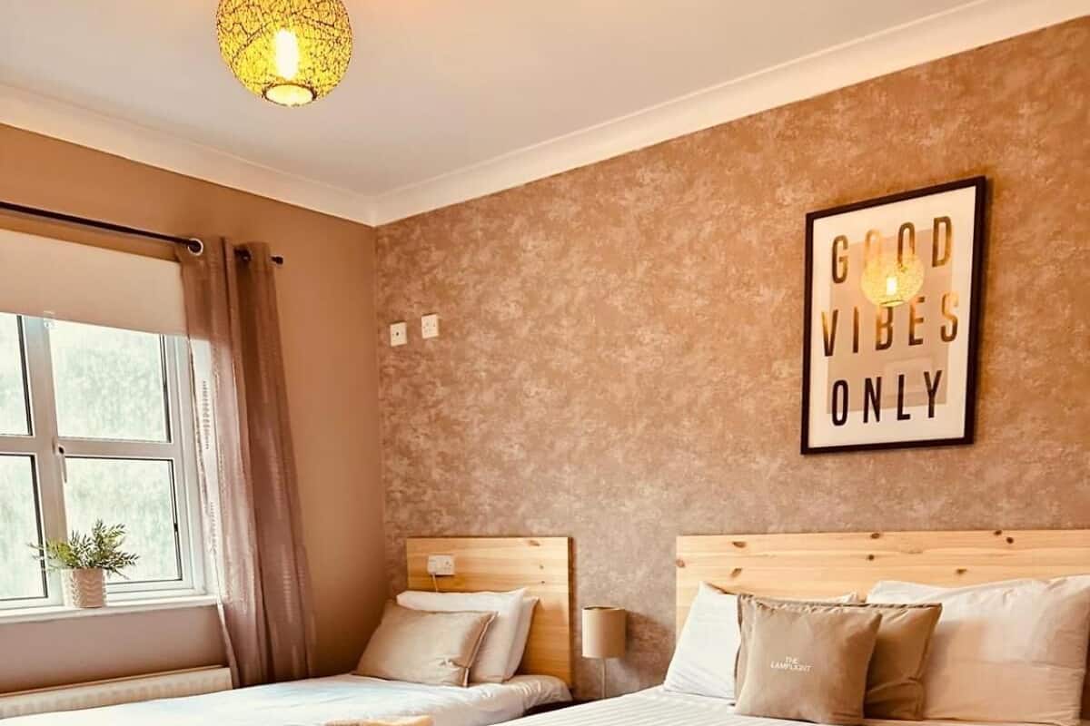 A hotel room with two beds, a window with curtains, a wall-mounted sign saying "GOOD VIBES ONLY", and a hanging light fixture.