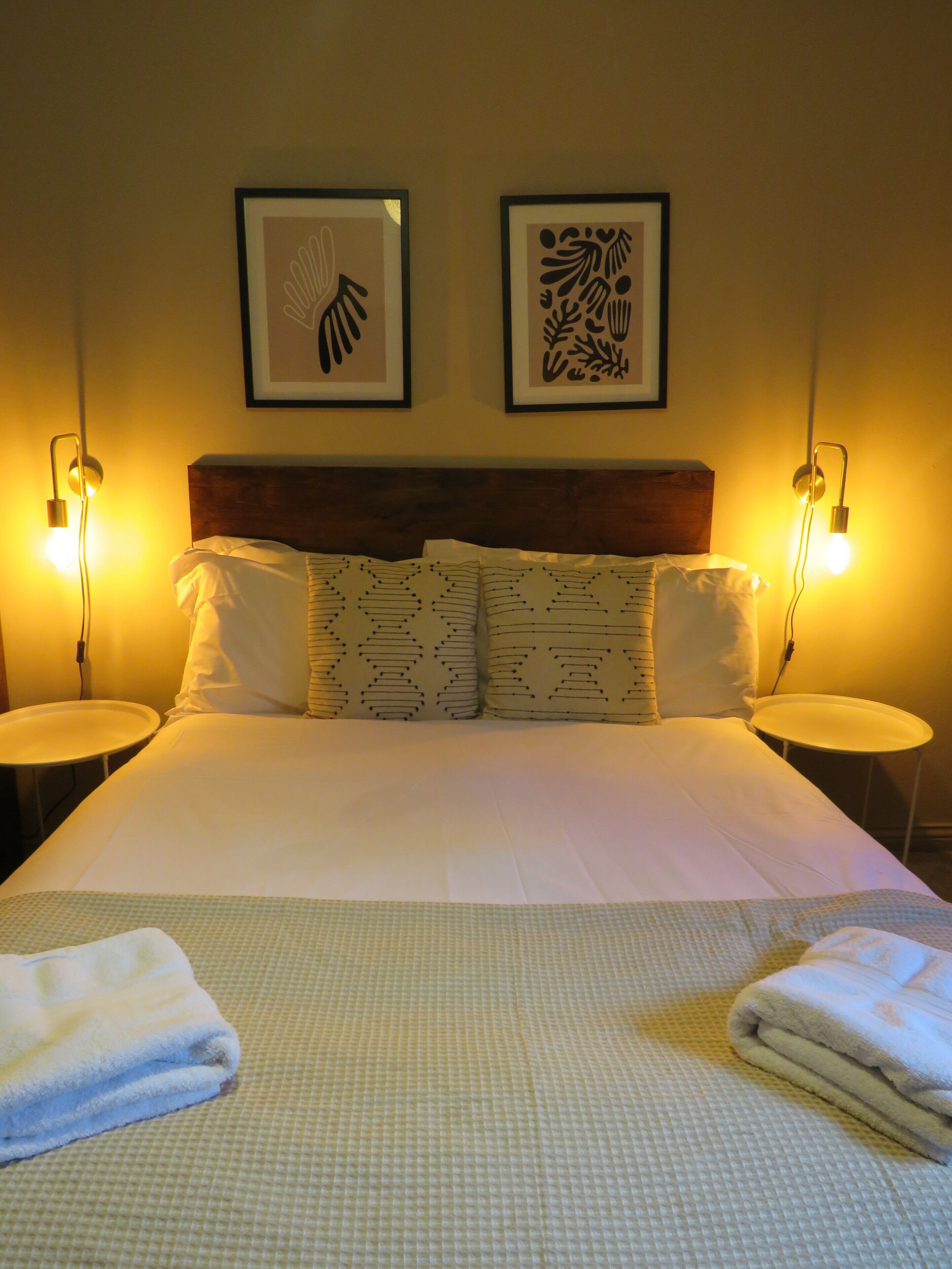 Une chambre d’hôtel avec un grand lit, deux tables de chevet, deux lampes fixées au mur et deux œuvres d’art encadrées au-dessus du lit.