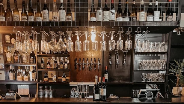 Zona de bar con tres sillas marrones, una barra con botellas y vasos, y una pared decorada con copas y botellas colgantes.