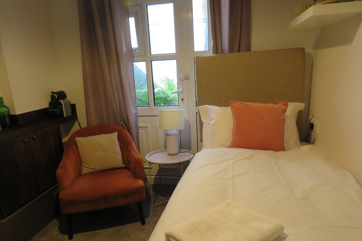 A hotel room with a bed, a chair, a small table with a lamp, and a window with curtains.