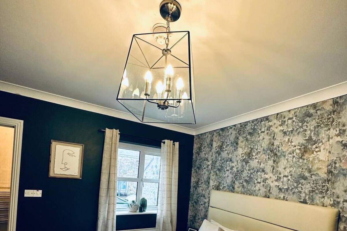 A neatly made bed with white linens and towels, a modern geometric chandelier, a window with curtains, and a textured wall.