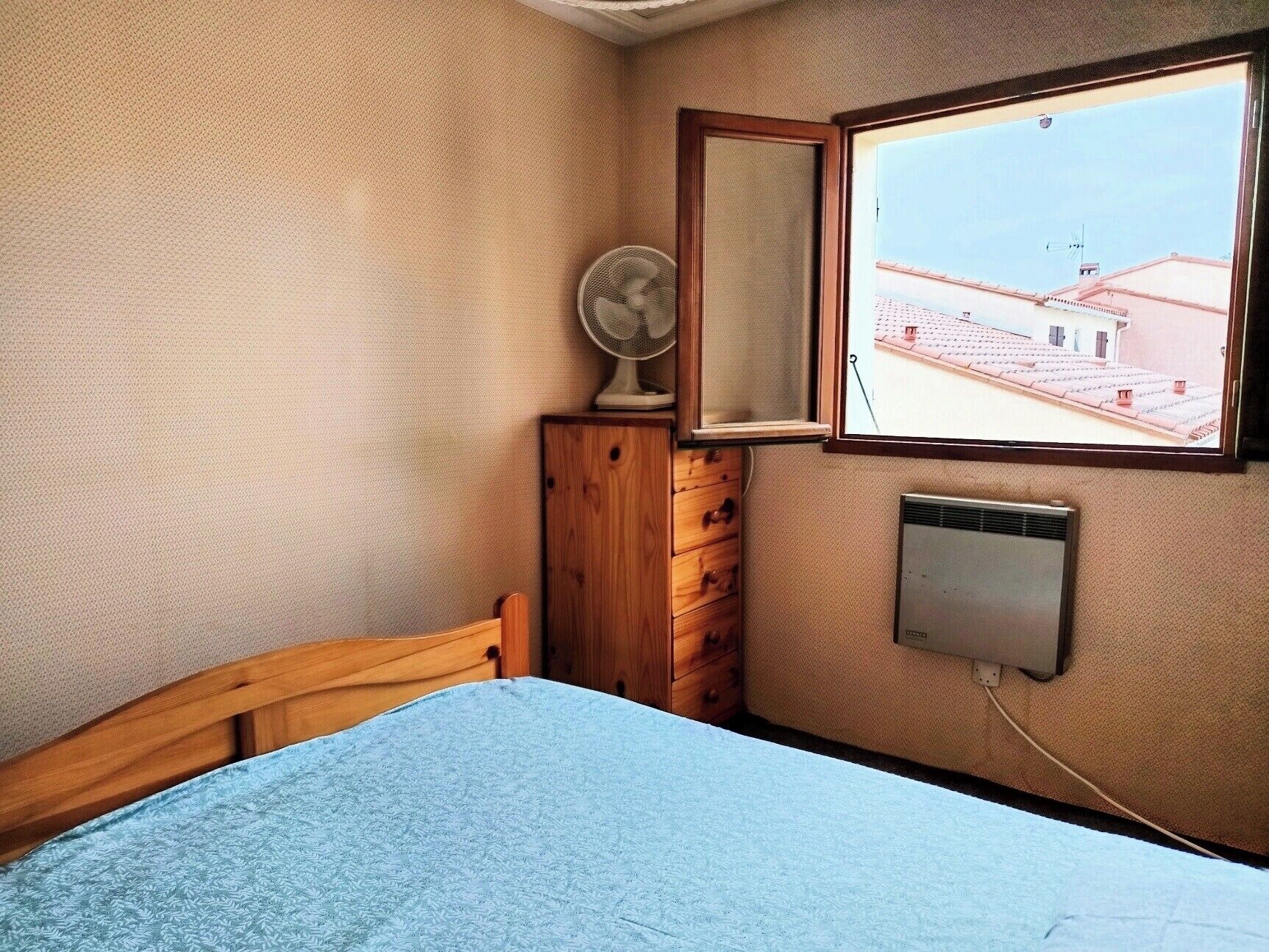 Ein Schlafzimmer mit einem Bett, einer Holzkommode, einem Ventilator, einem Fenster mit Blick auf Gebäude und einem Wandheizgerät.