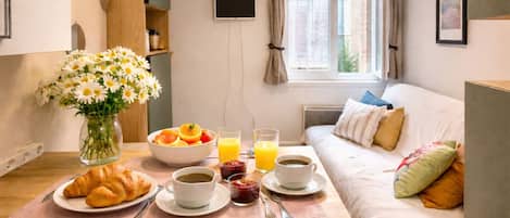 A breakfast setup with croissants, coffee, juice, and a vase of flowers on a wooden table in a room with a bed and a window.