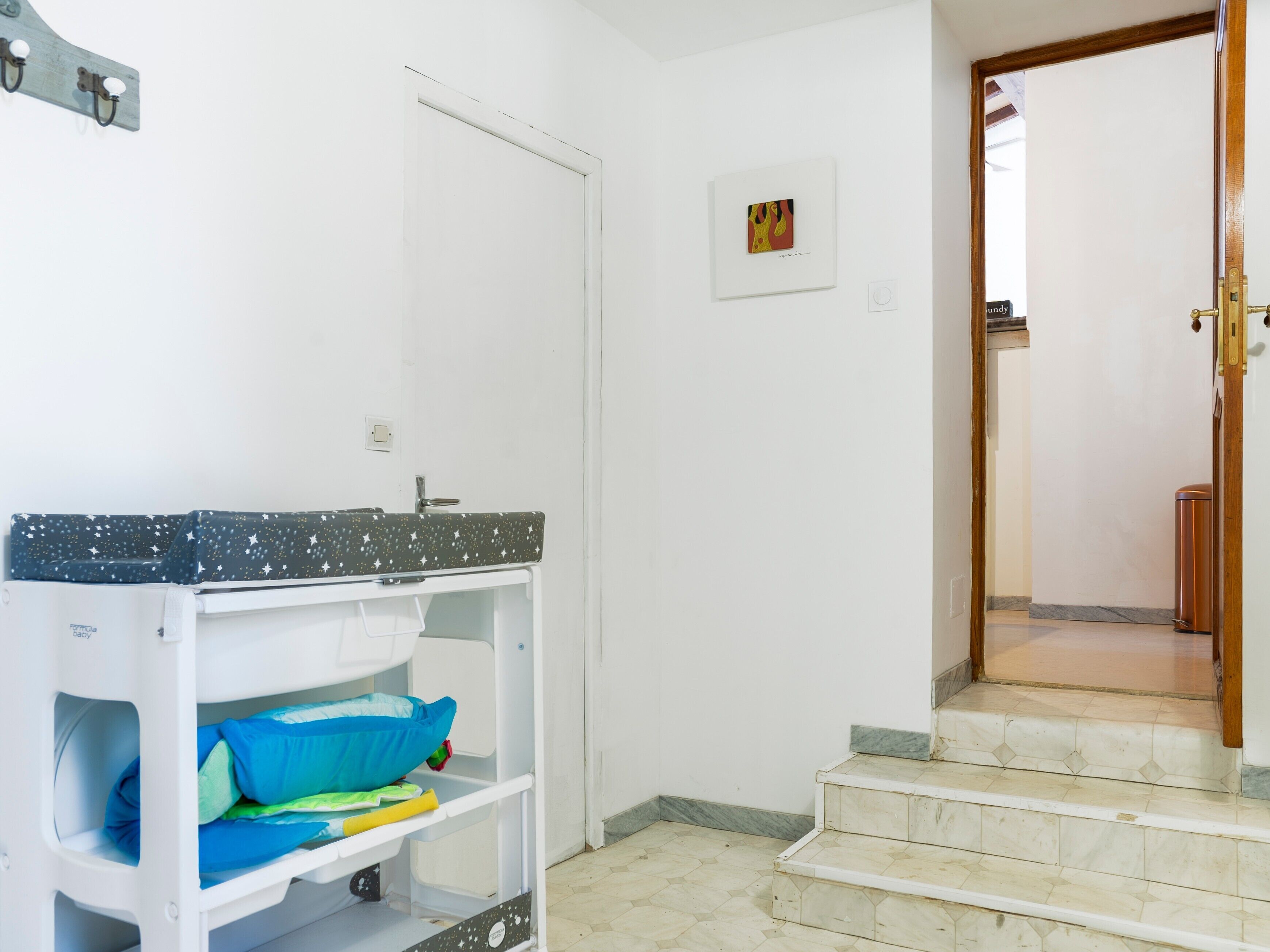 A room with a white door, a small framed picture on the wall, a white shelf with a star-patterned top, and a set of stairs leading to another room.