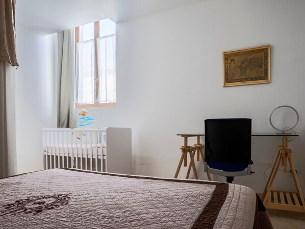A room with a crib, a desk with a chair, a large bed, a television on a stand, and a framed map on the wall.