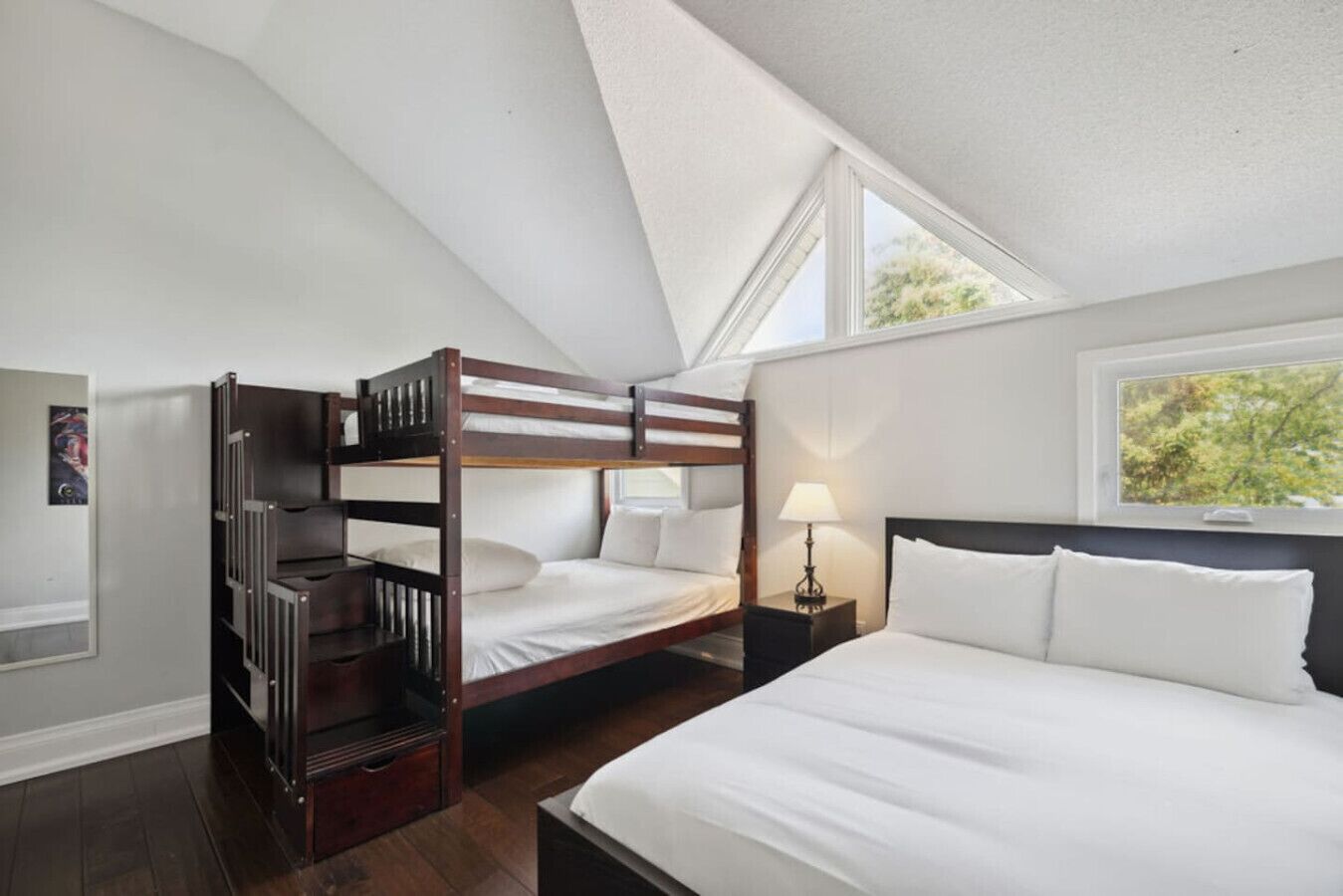 A room with a bunk bed, a single bed, a nightstand with a lamp, and a window with a view of trees.