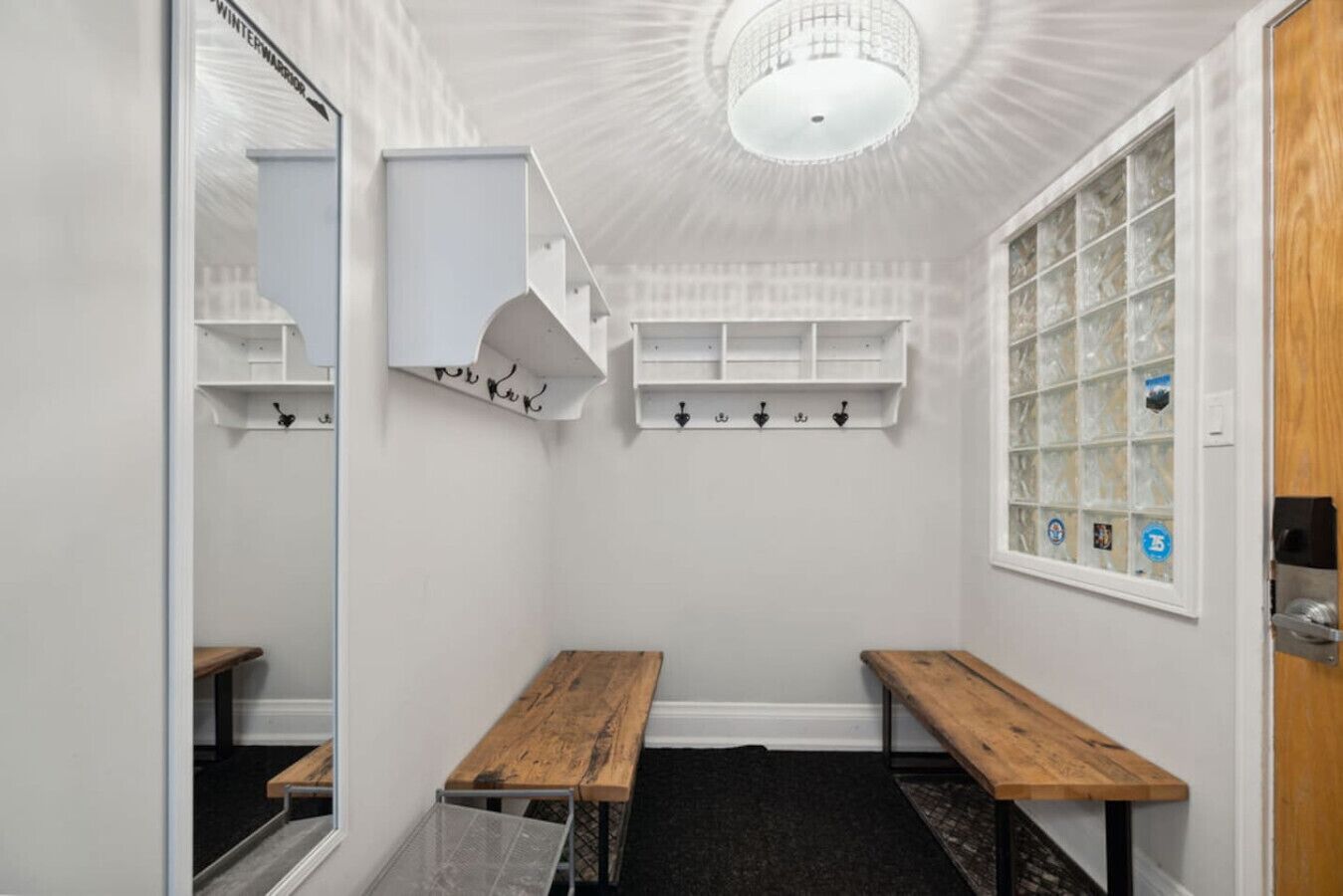 Une petite pièce aux dimensions réduites, avec des murs blancs, un banc en bois, un miroir et une fenêtre à carreaux.