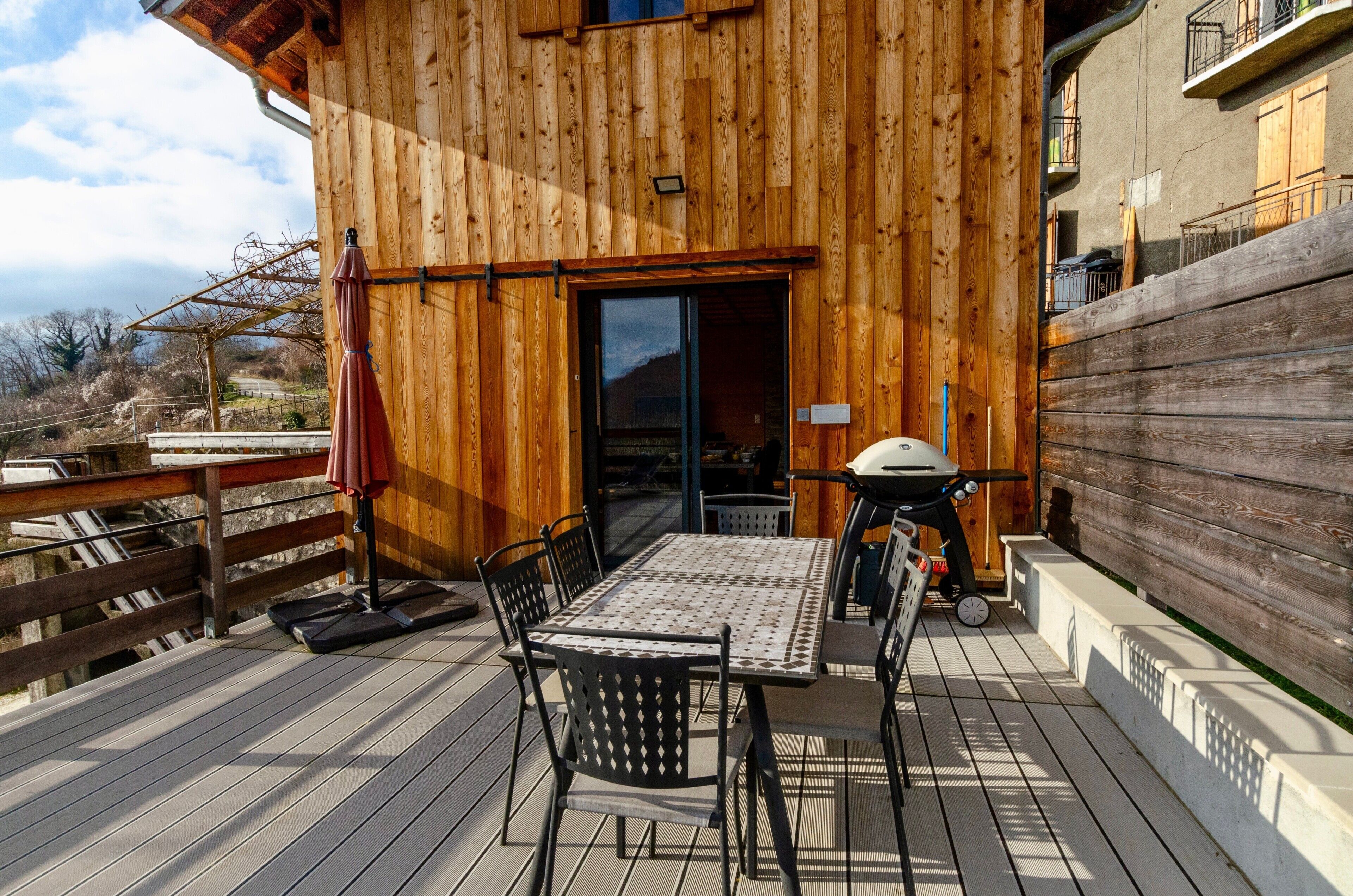 En træterrasse med et bord, stole, grill og udsigt over det omkringliggende landskab.