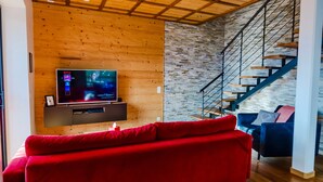 A modern living room with a TV, red and blue sofas, a wooden ceiling, and a staircase with a stone wall. - Chalet 'Au Pied De L'arcluse' with Mountain Views, Wi-Fi, and Air Conditioning (Saint-Pierre-d'Albigny)