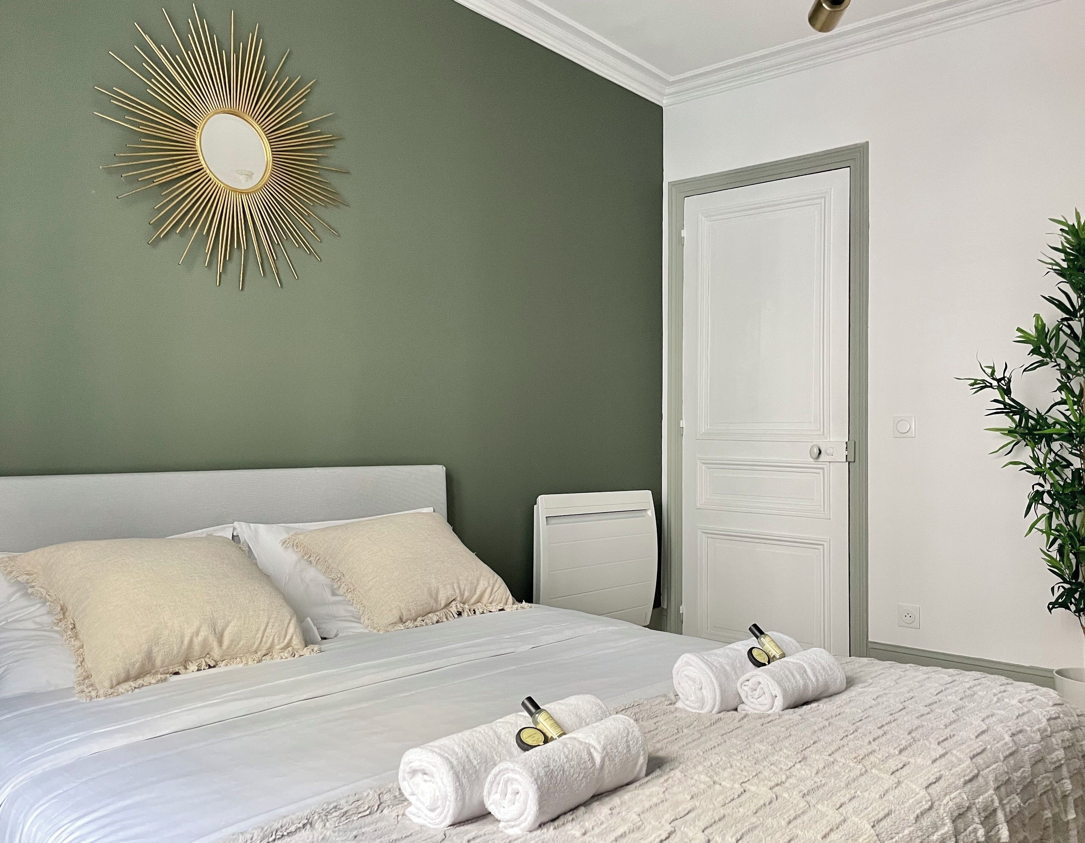 A neatly made bed with white linens, a gold sunburst mirror on the wall, and a white door with a plant beside it.