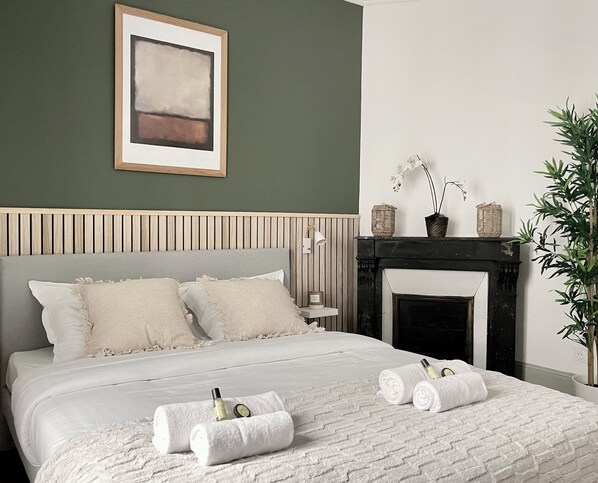 A neatly made bed with pillows, a bedside table with a lamp, a framed abstract painting, a fireplace, and a potted plant. - 1590 Legendre (Paris)