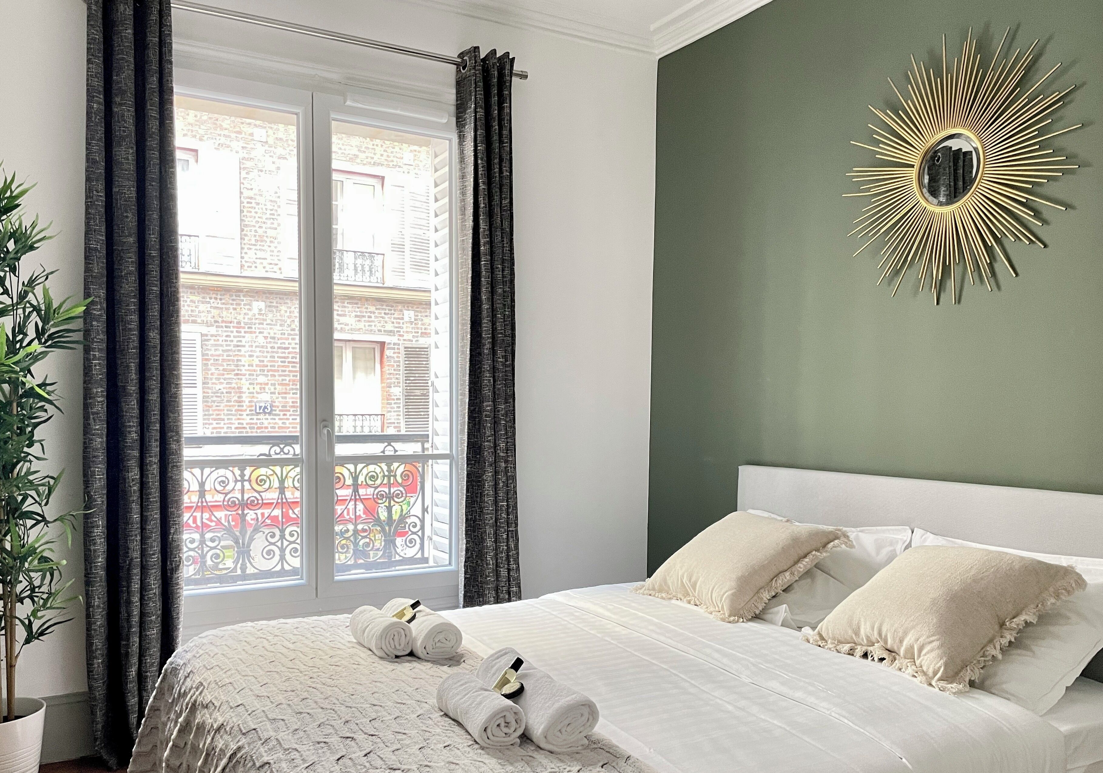 A bedroom with a bed, a window with curtains, a potted plant, and a wall-mounted mirror.