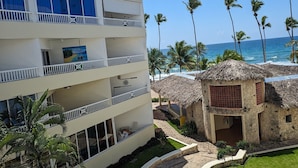 Un hôtel à plusieurs étages doté de balcons donnant sur une plage, des palmiers et une structure surmontée d’un toit de chaume.