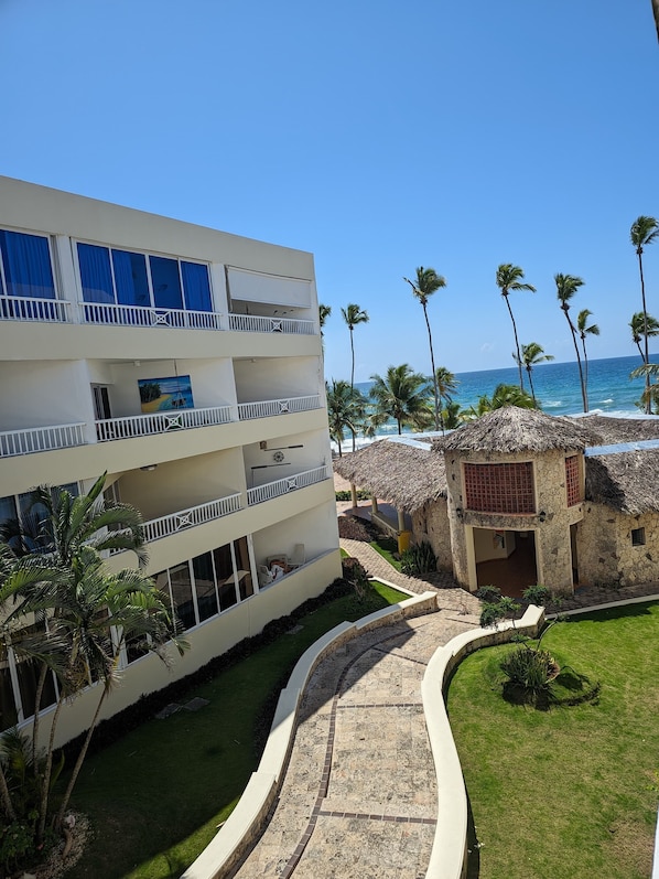 A multi-story hotel with balconies overlooking a beach, palm trees, and a thatched-roof structure.