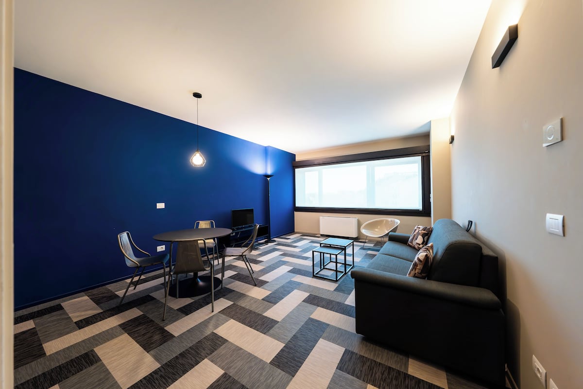 A modern living room with a blue accent wall, a black sofa, a coffee table, and a dining area with chairs and a small table.