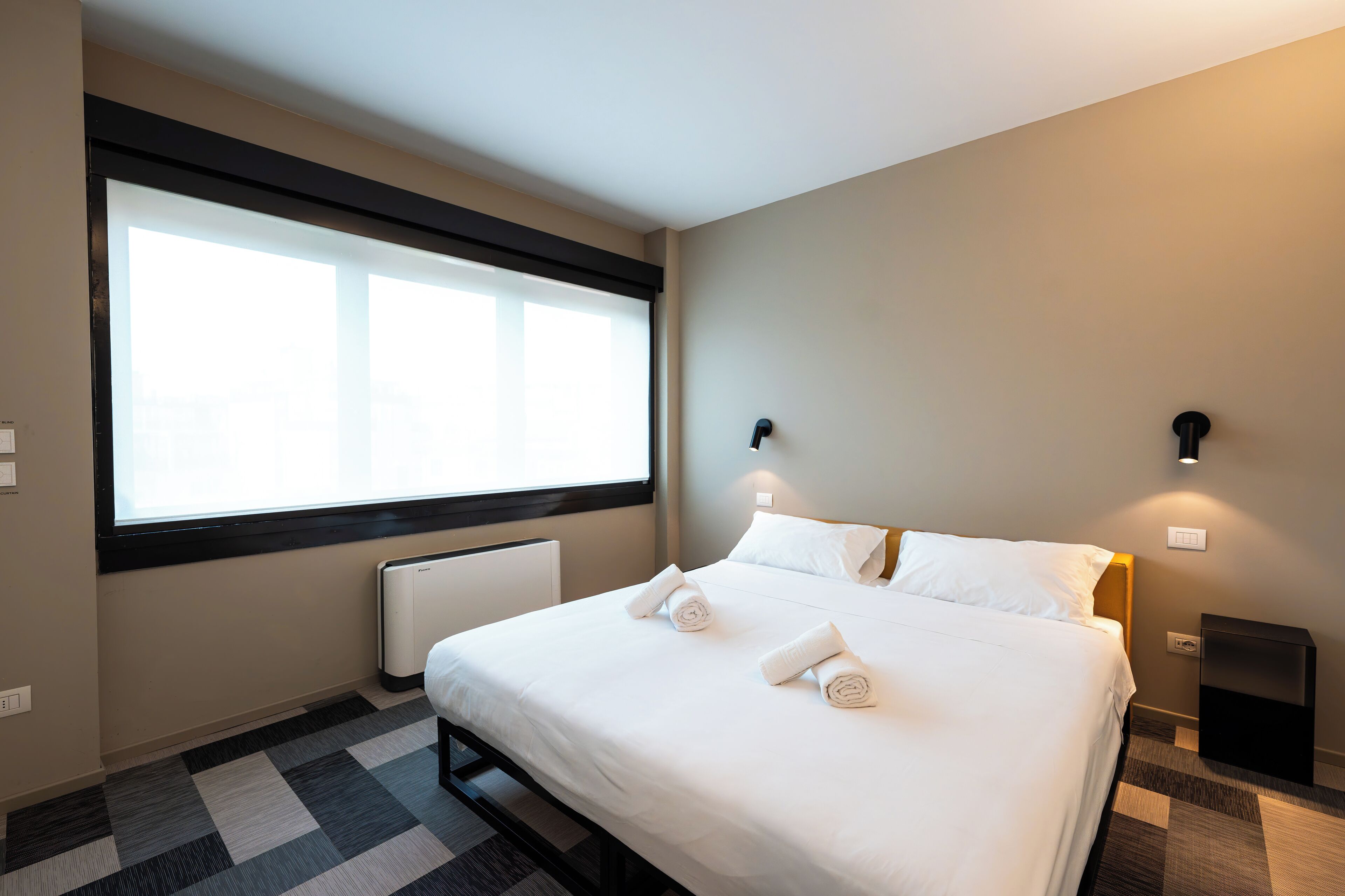 A hotel room with a large window, a bed with white linens, two wall-mounted lights, and a black nightstand.