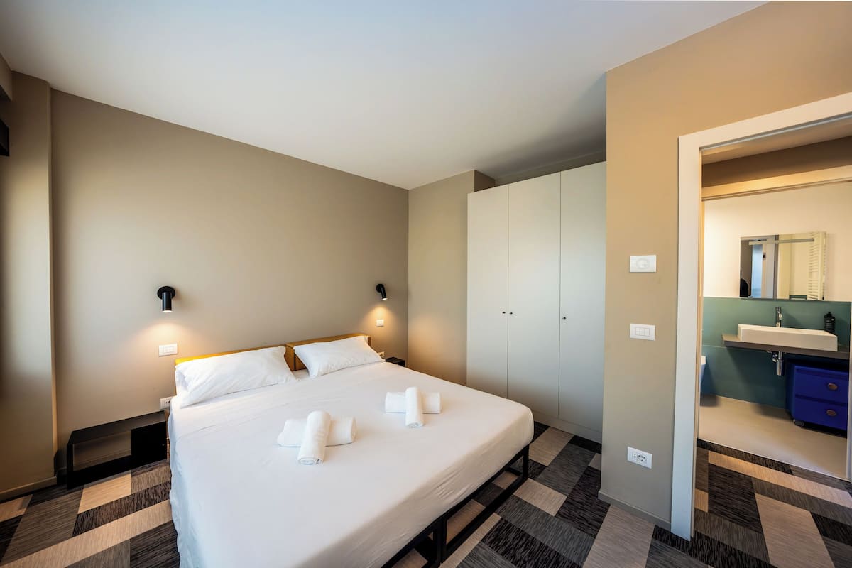 A hotel room with a bed, bedside lamps, a wardrobe, and a bathroom with a sink and mirror.