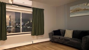 A room with a window overlooking a cityscape at night, a gray sofa, and a painting on the wall. - Incredible NYC and Water Views 3 bedrooms only 15 min from NYC New Floors  (West New York)
