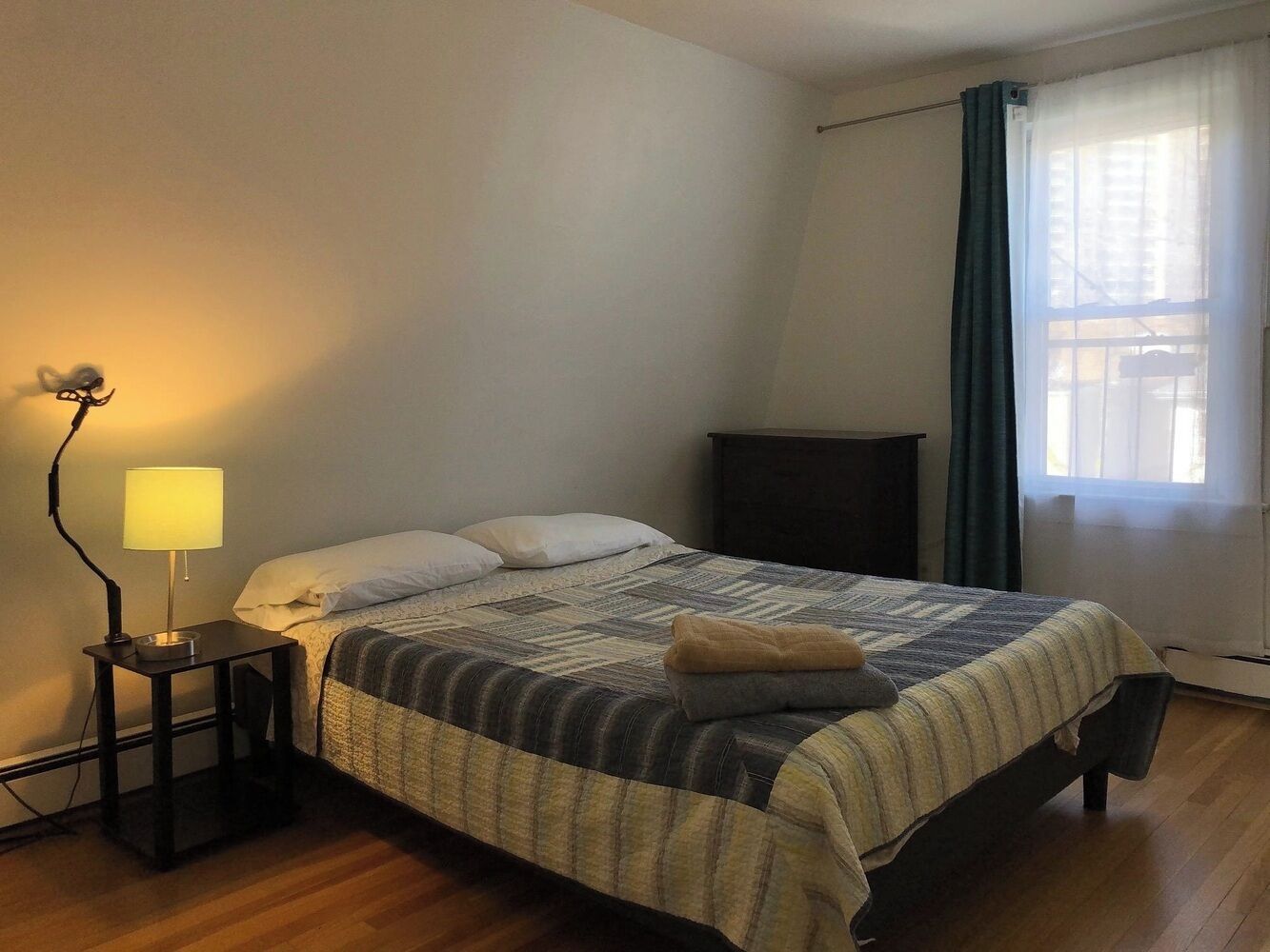 Un dormitorio con cama, lámpara de noche, cómoda y ventana con cortinas.