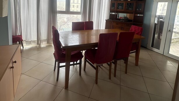 A dining area with a wooden table and red chairs, a cabinet, and a view of a building through large windows.