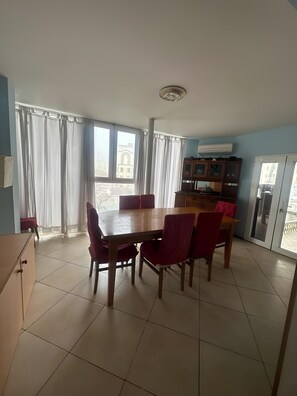 Een eetgedeelte met een houten tafel en rode stoelen, een kast en uitzicht op een gebouw door grote ramen.