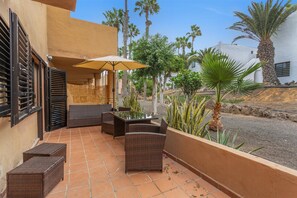Une terrasse extérieure avec des meubles en osier, une table à plateau en verre et un parasol beige.