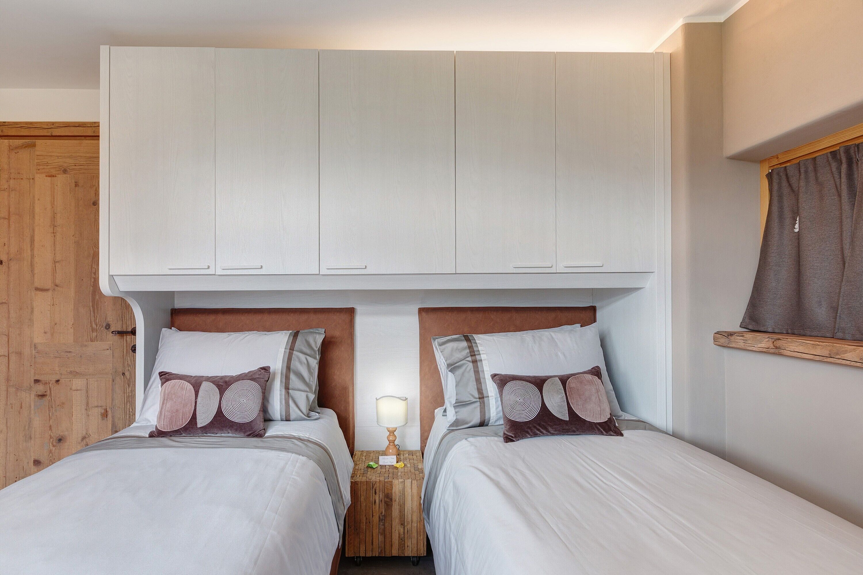 Une chambre avec deux lits, une tête de lit en bois, une table de chevet avec une lampe et une armoire placée au-dessus des lits.