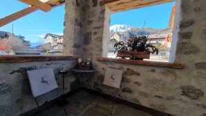 Un intérieur rustique avec un mur en pierre, des poutres en bois, une fenêtre ornée d’une plante et deux tableaux encadrés.