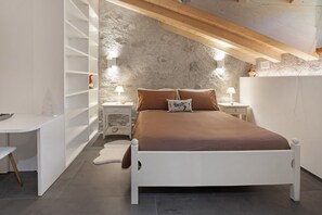 Ett sovrum med en vit säng, ett träfärgat sänggavel och en vägg av sten. Det finns två sängbord med lampor, en vit garderob och ett vitt skrivbord med en stol.