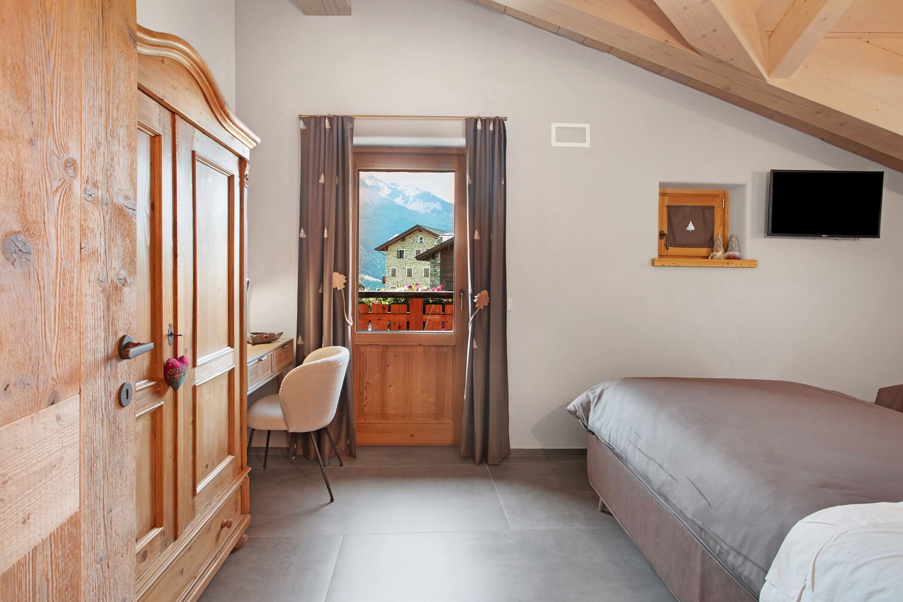 A bedroom with a bed, a wooden wardrobe, a chair, a television, and a window with a view of a mountain.